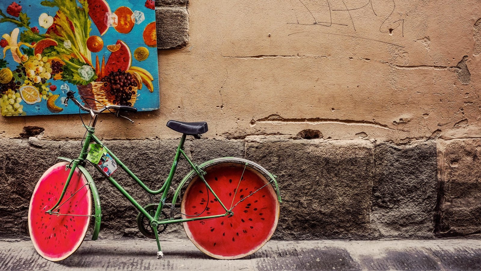 Green bicycle with watermelon-designed wheels, promoted by AI Marketing, leaning against a weathered wall next to a colorful painting.